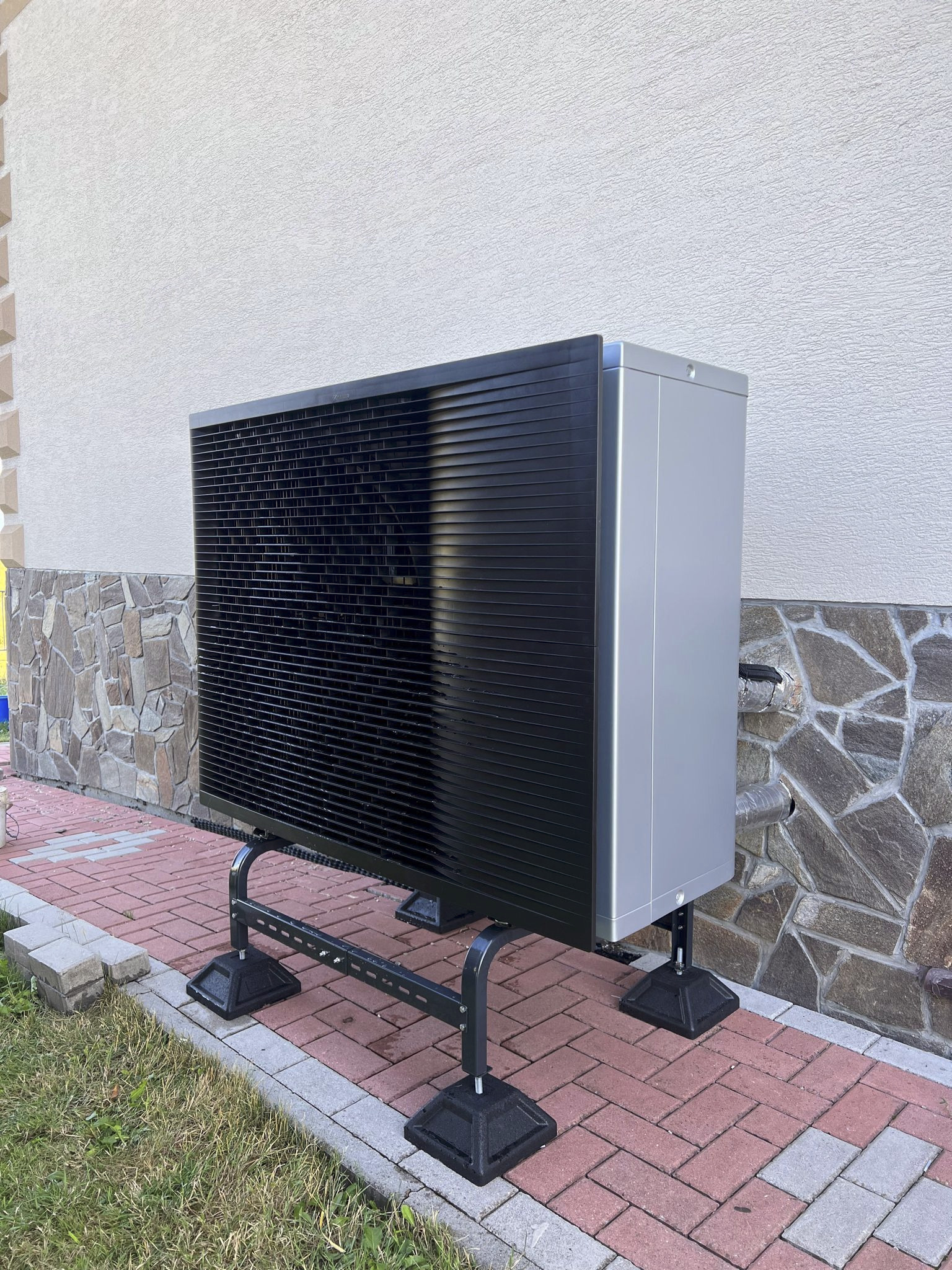 Zewnętrzna jednostka pompy ciepła powietrze-woda, ustawiona na metalowej ramie, z widocznymi przyłączami. Tło stanowi ściana budynku z elementami kamiennej elewacji.