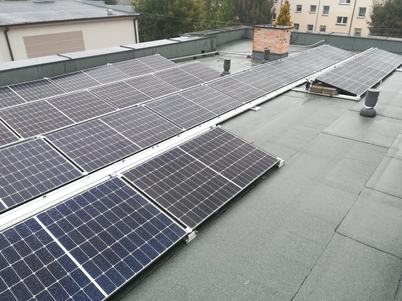 Widok z góry na instalację fotowoltaiczną na płaskim dachu budynku. Panele w rzędach, z widocznymi elementami montażowymi. Dach pokryty papą. W tle budynki i drzewa.