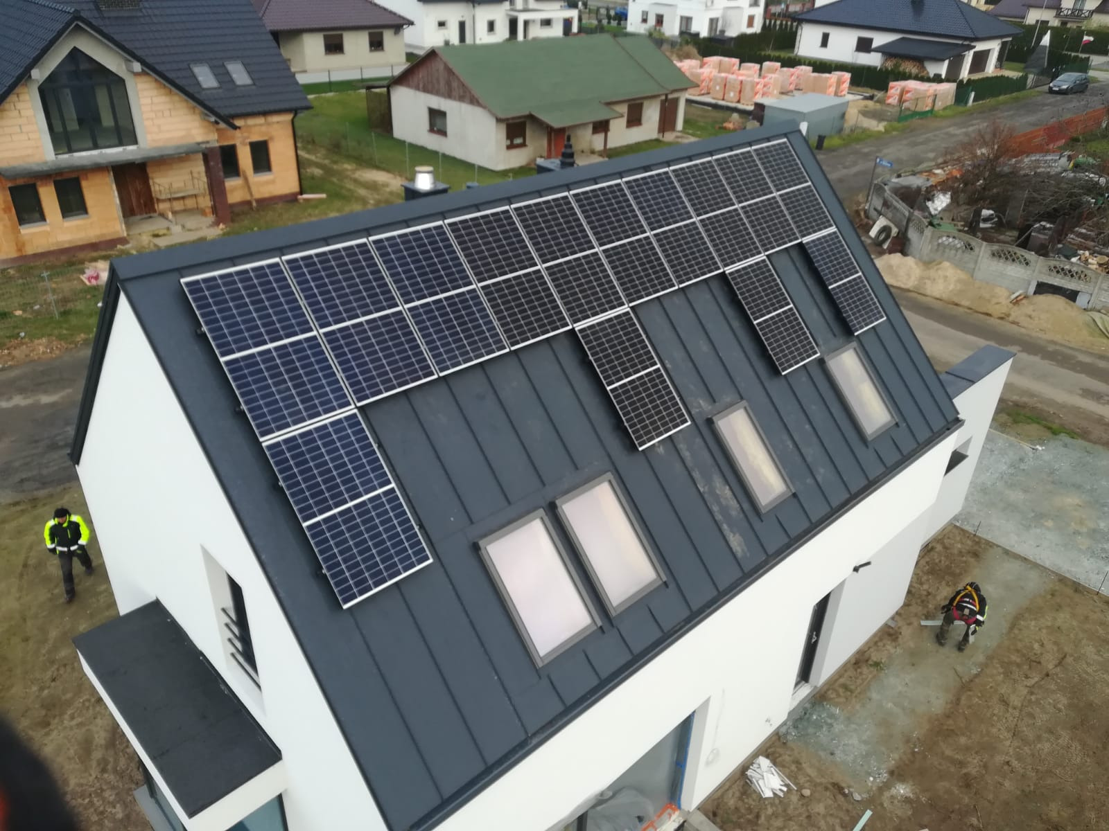 Fotowoltaika na dachu domu jednorodzinnego z szarej blachy, widok z góry na panele słoneczne i okna dachowe. Na dole widoczni pracownicy w odblaskowych ubraniach.