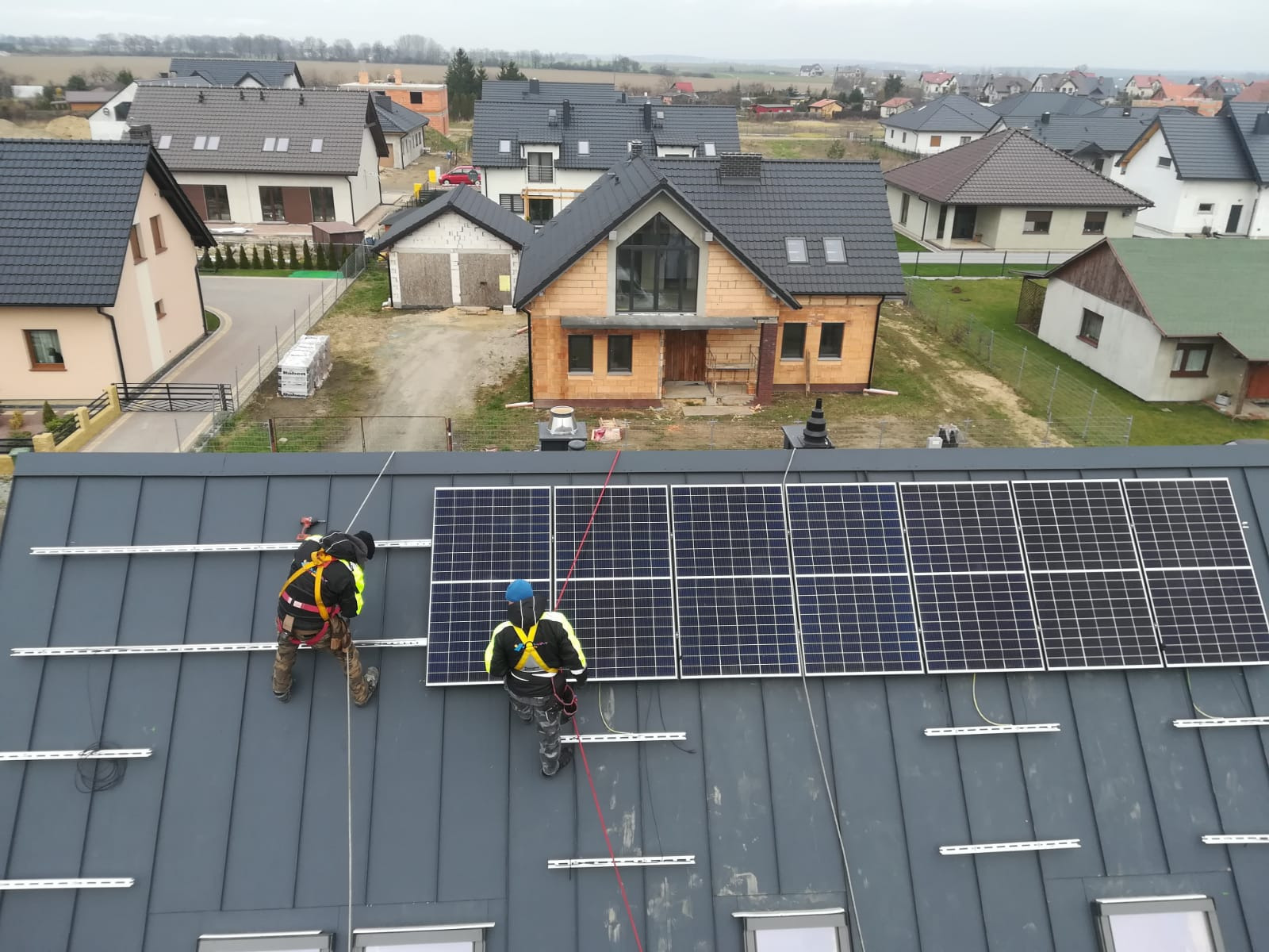 Montaż paneli fotowoltaicznych na szarym dachu domu jednorodzinnego przez dwóch pracowników w uprzężach, z widokiem na okoliczne budynki.