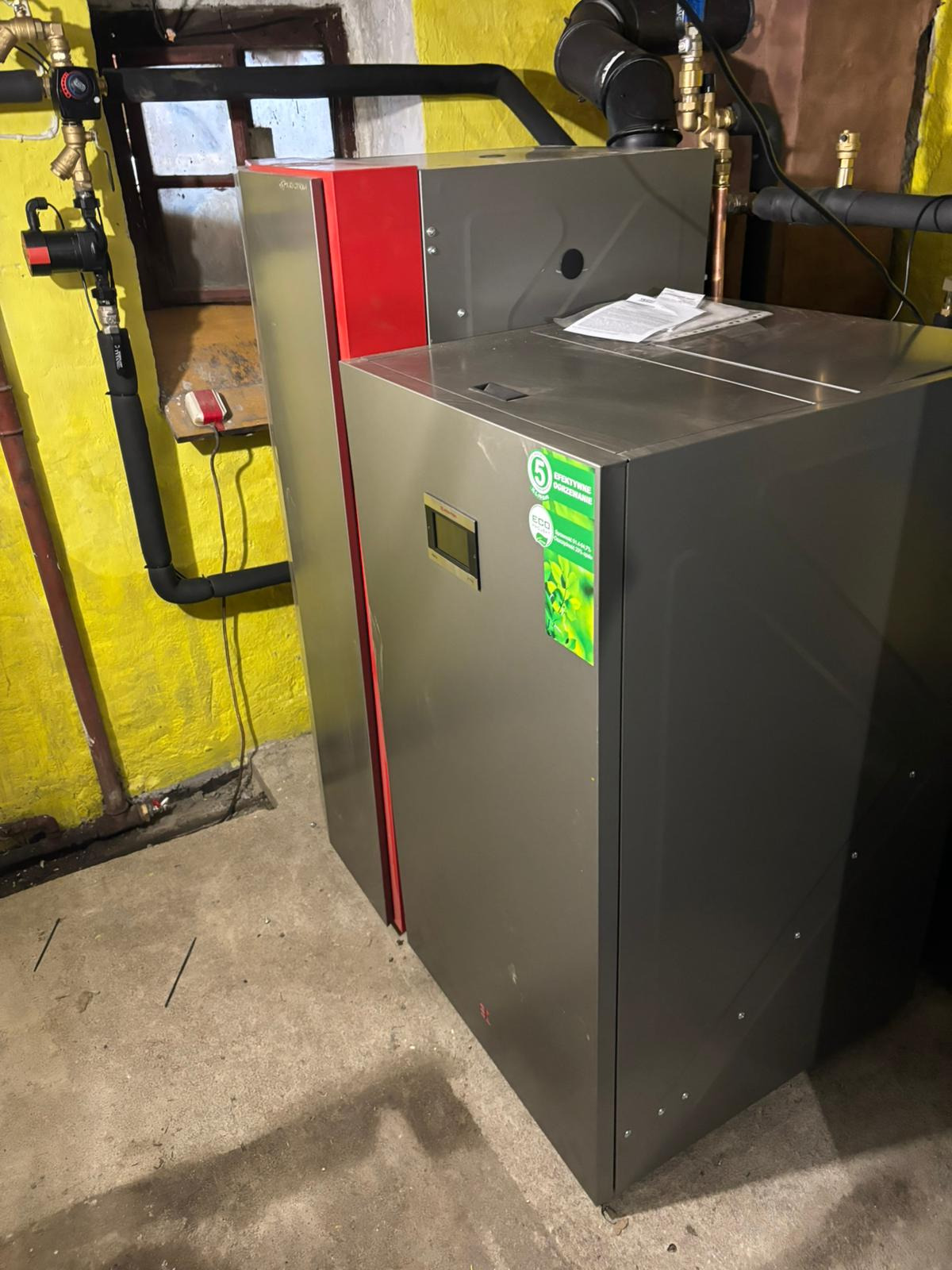 Nowoczesny, szary piec CO z czerwonym akcentem, zainstalowany w kotłowni o żółtych ścianach i betonowej podłodze. Widoczne rury instalacji grzewczej.