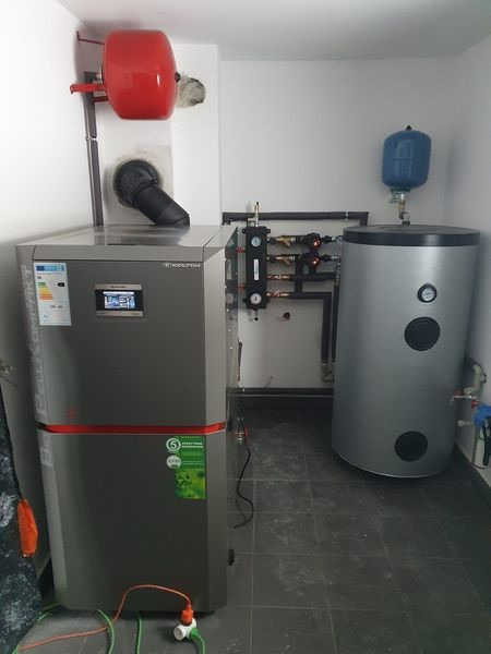 Szary piec z czerwonym zbiornikiem i srebrny zasobnik z niebieskim naczyniem wzbiorczym w kotłowni na ciemnej podłodze. Widoczne rury i elementy instalacji grzewczej.