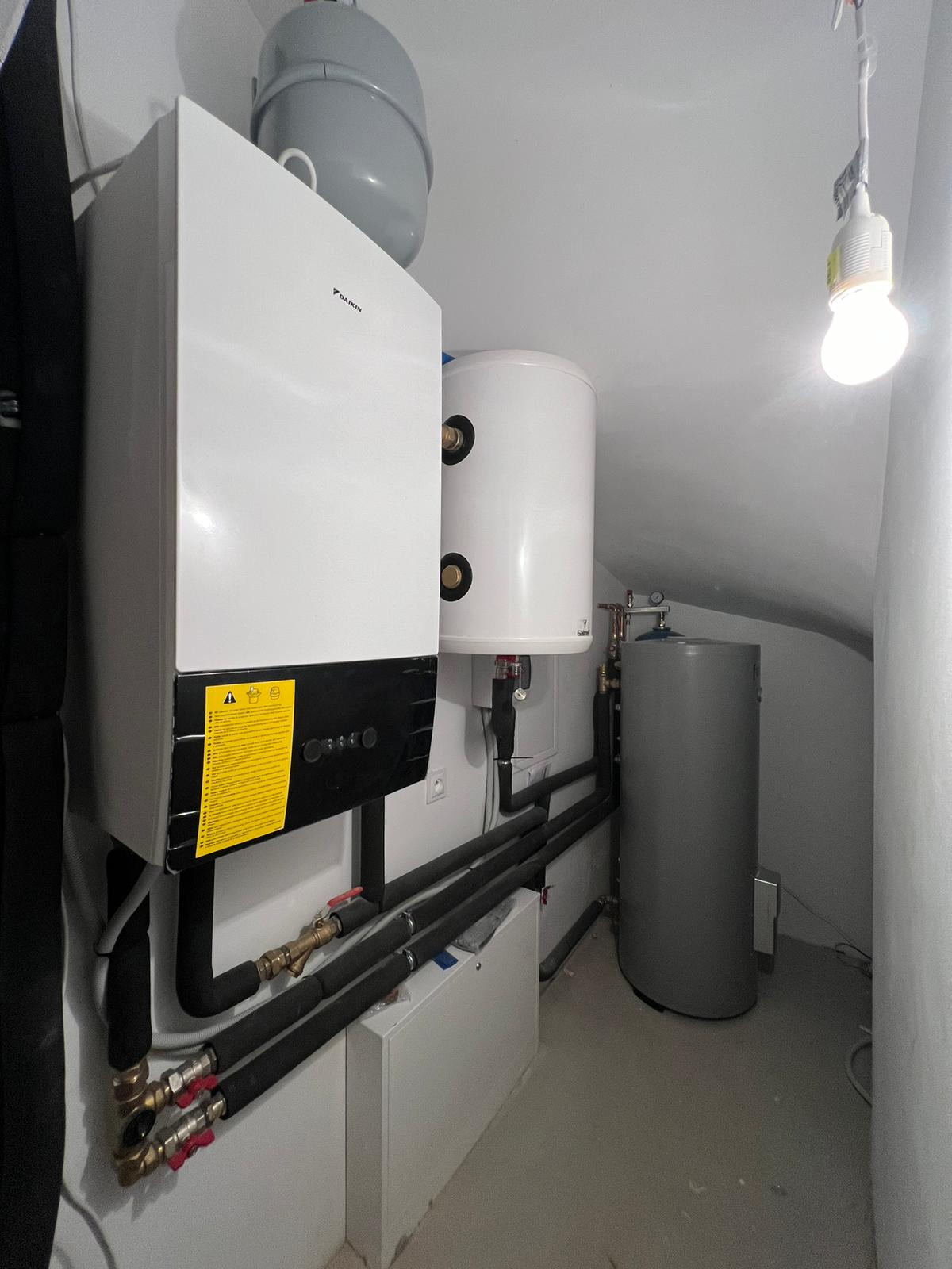 Biały piec grzewczy Daikin z żółtą naklejką, zbiornik wyrównawczy i dwa zasobniki ciepłej wody w kotłowni. Widoczne rury i zawory.