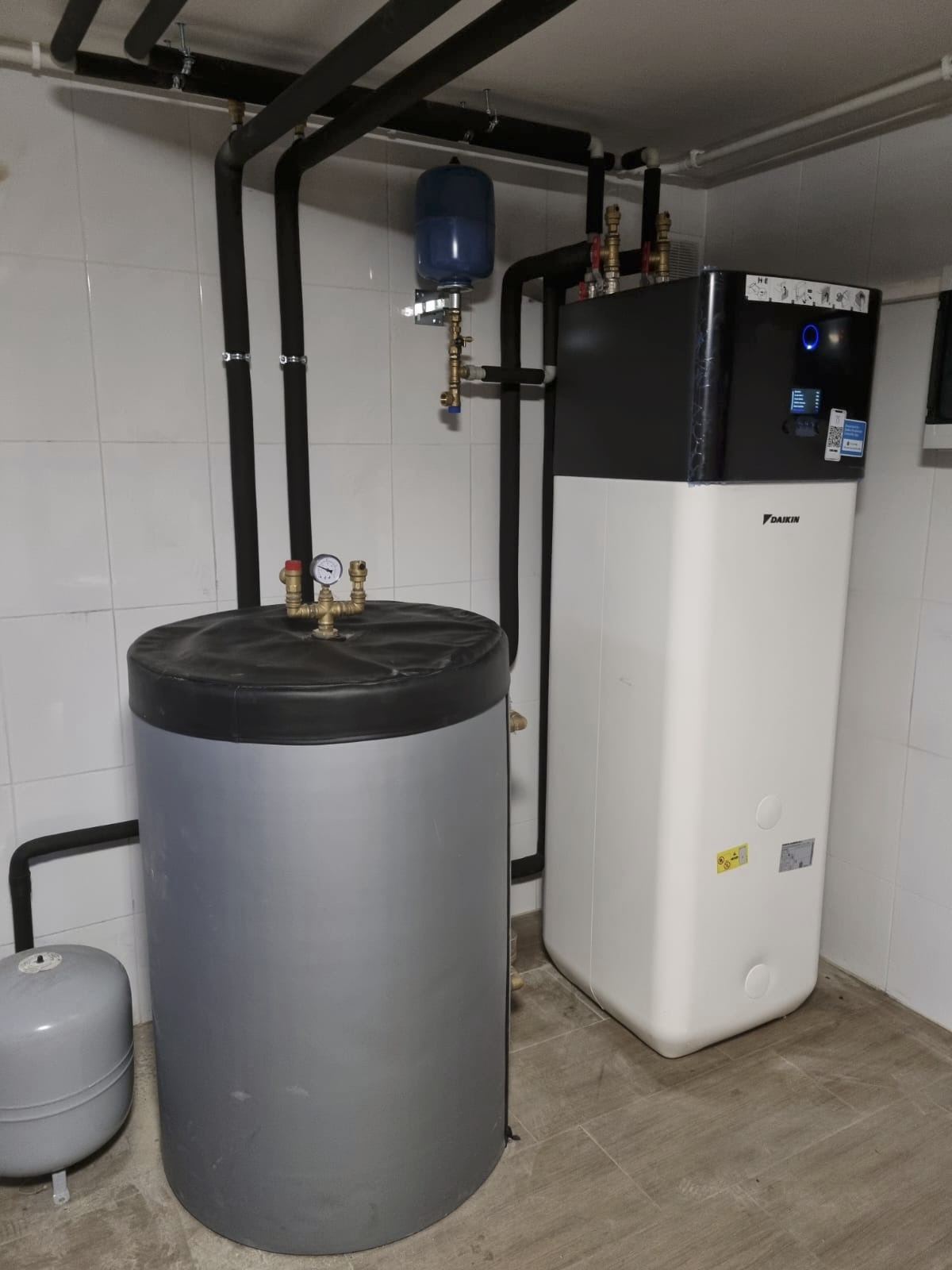 Zestaw grzewczy Daikin z zbiornikiem i rurami w kotłowni wyłożonej białymi płytkami. Widoczne manometry i zawory, instalacja pod sufitem.