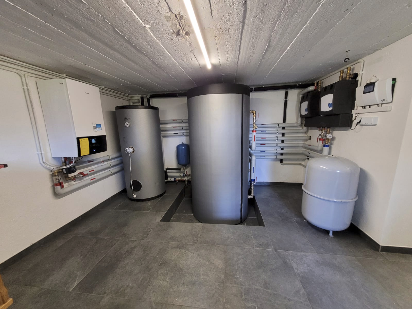 Kotłownia z dwoma srebrnymi zbiornikami akumulacyjnymi, białym piecem i instalacją rurową z zaworami na ścianach. Podłoga wyłożona szarymi płytkami.