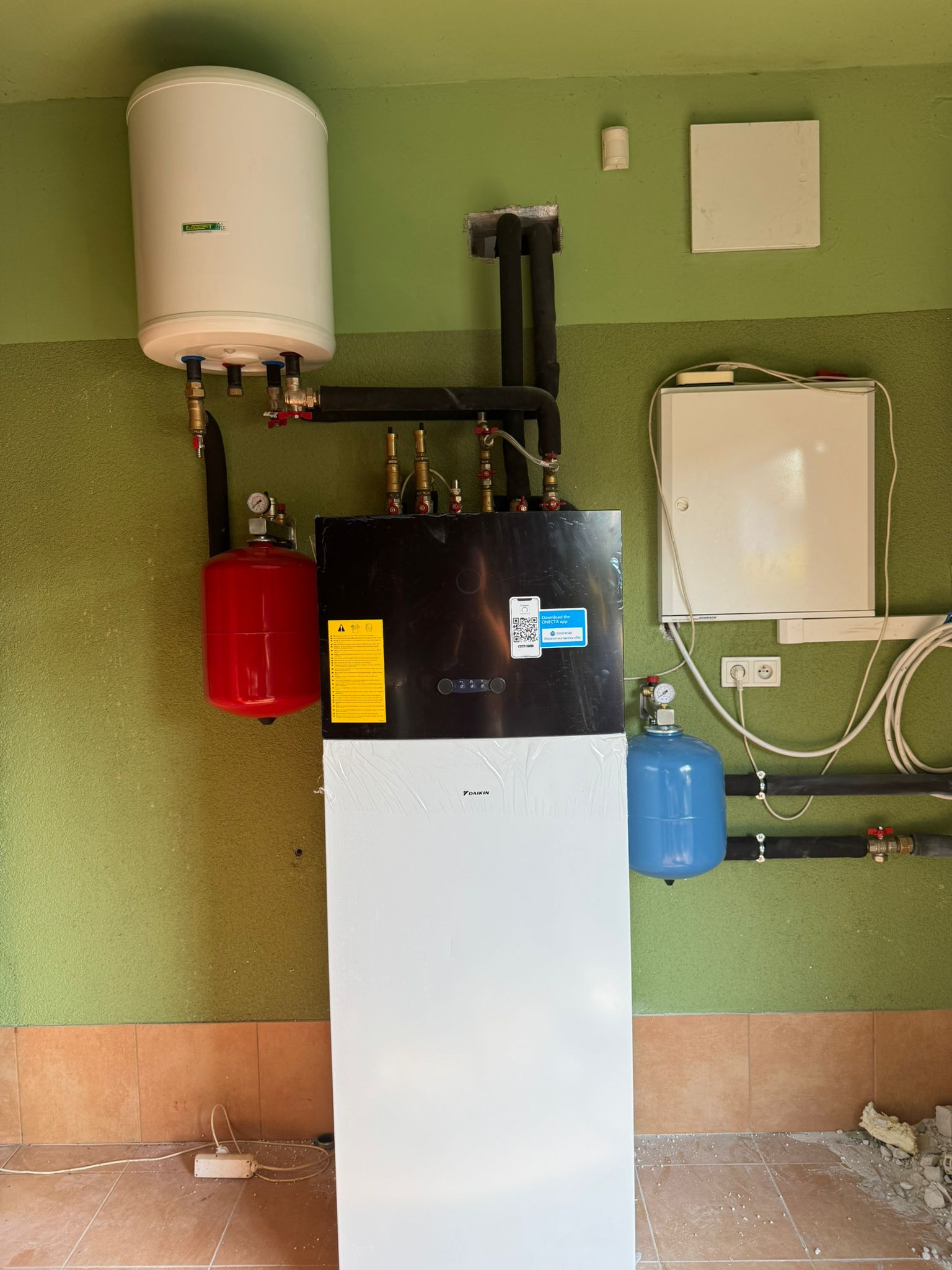 Nowoczesny piec CO Daikin z zbiornikiem, czerwonym i niebieskim naczyniem przeponowym na tle zielonej ściany i brązowych płytek podłogowych.