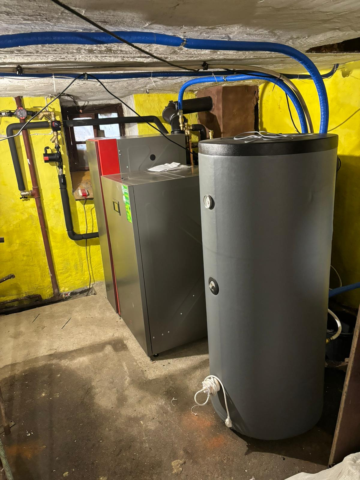 Szary piec CO z czerwonym panelem bocznym i bojler w piwnicy z żółtymi ścianami, widoczne rury instalacyjne i okno.