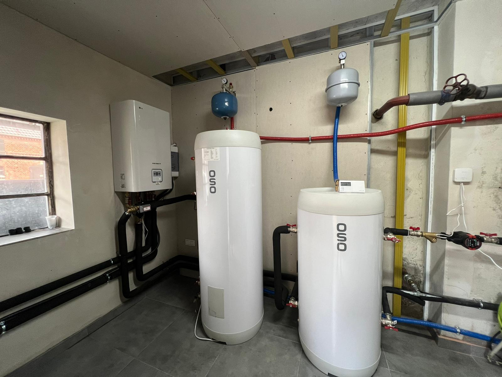 Dwa białe zbiorniki OSO z podłączeniami hydraulicznymi w kotłowni z szarą podłogą, widoczny piec LG i instalacja rurowa. Ściany częściowo wykończone płytami.
