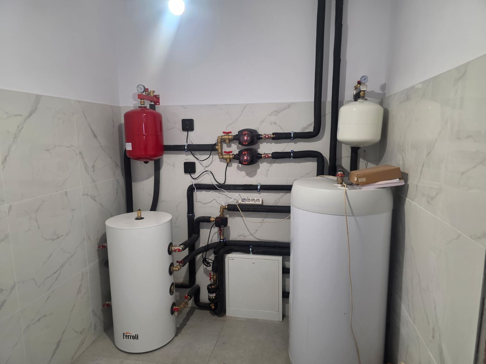Zainstalowana kotłownia w pomieszczeniu z marmurowymi płytkami. Widoczne dwa białe zbiorniki Ferolli, czerwony zbiornik wyrównawczy i czarne rury.