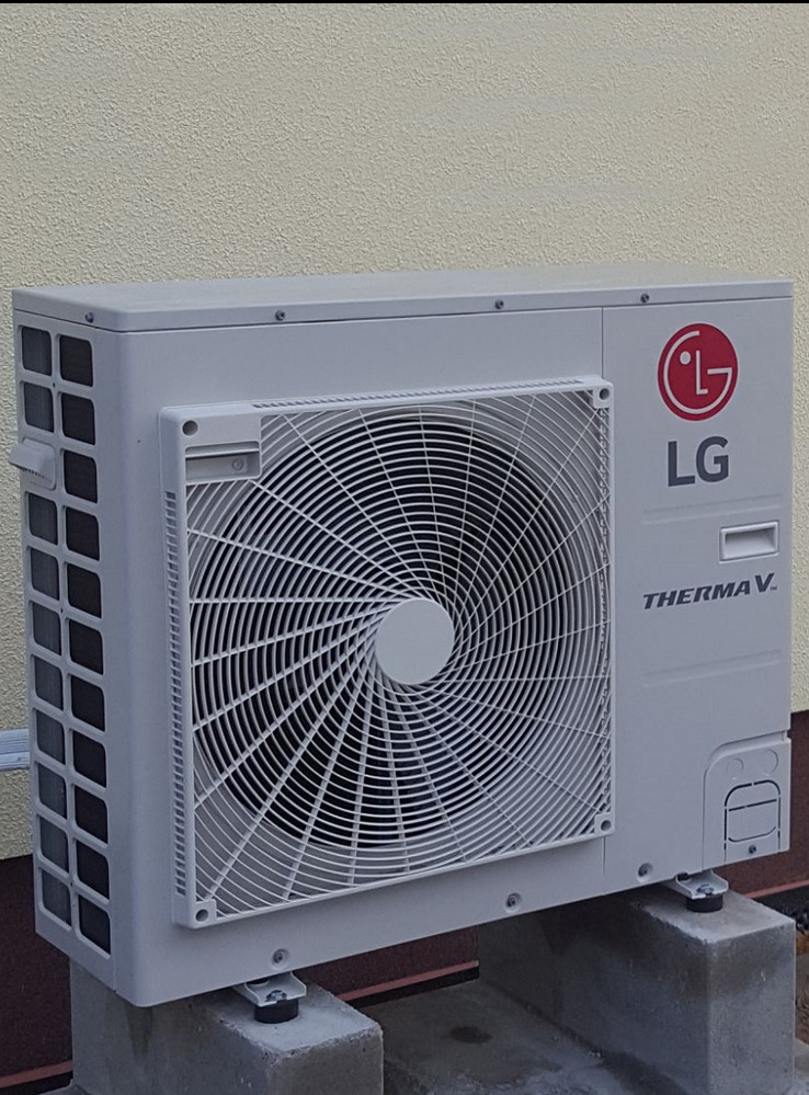 Zewnętrzna jednostka pompy ciepła LG Therma V na betonowych podstawach, przylegająca do ściany budynku o jasnej elewacji. Widoczny wentylator i logo producenta.