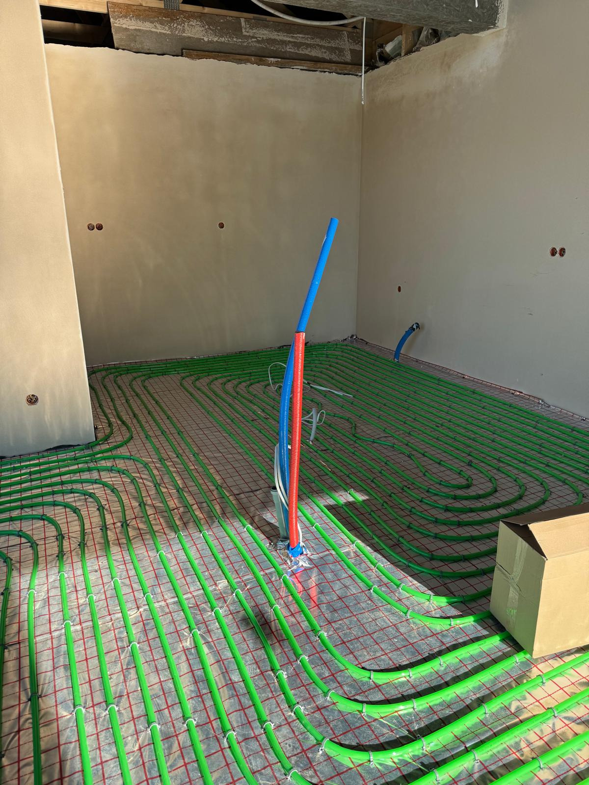Zielone rurki ogrzewania podłogowego ułożone na czerwonej siatce w surowym wnętrzu. Pionowe rury niebieskie i czerwone wystają z podłoża. Widoczny karton.