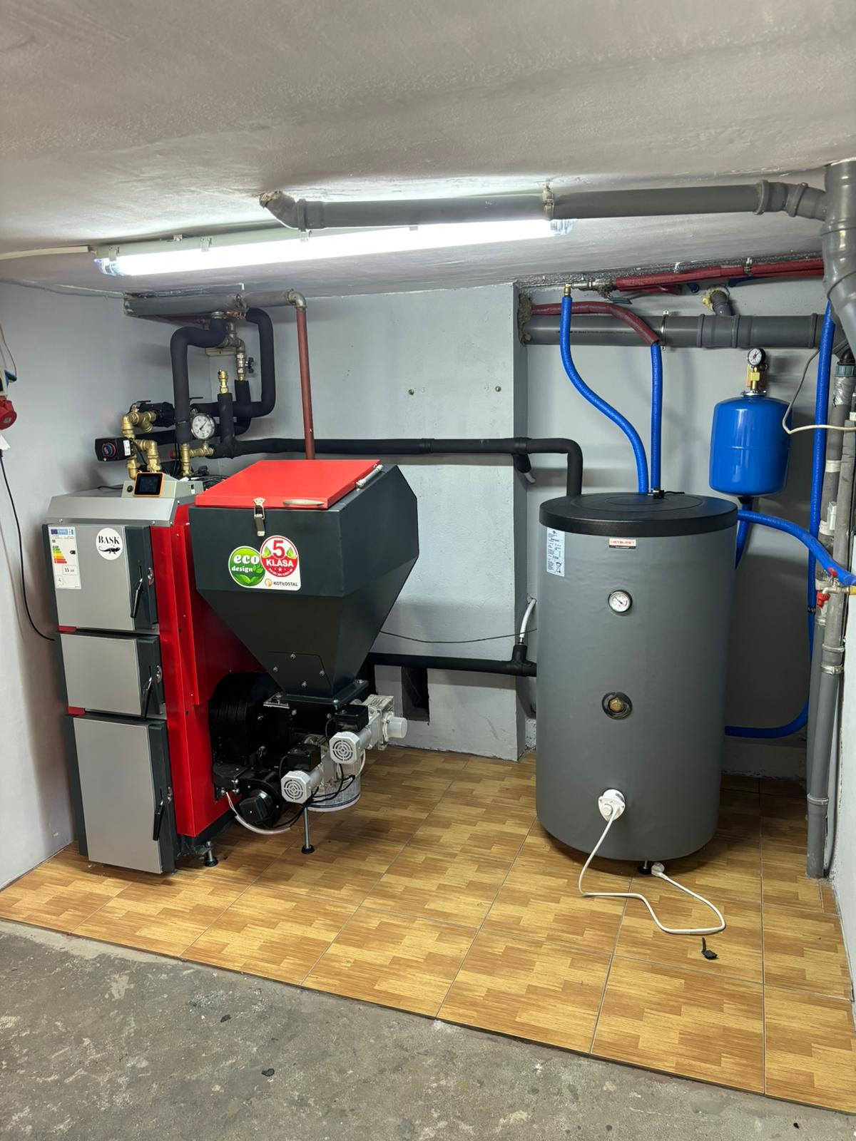 Kotłownia z nowoczesnym piecem na ekogroszek z podajnikiem i zasobnikiem, oraz zbiornikiem wyrównawczym. Widoczne rury i instalacja.
