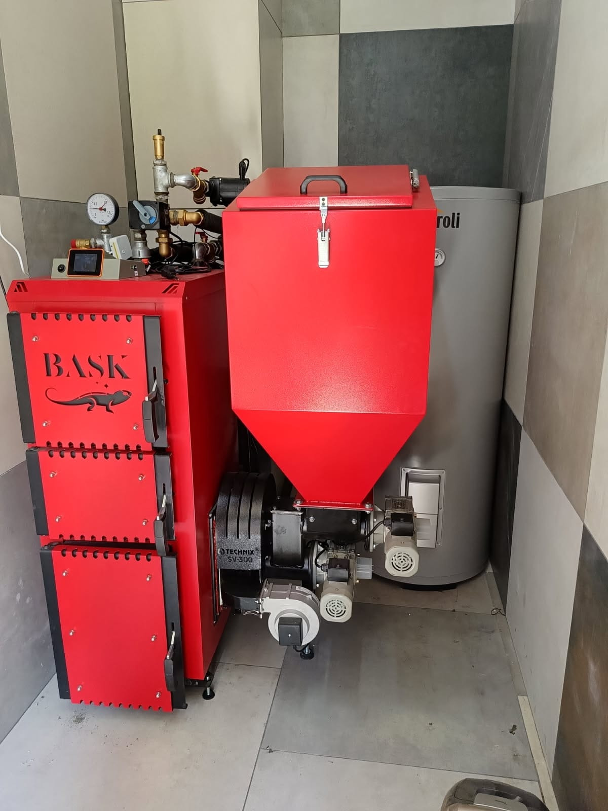 Czerwony piec CO marki BASK z podajnikiem na ekogroszek, model TECHNIX SV-500, obok szary zasobnik na wodę marki Roli w kotłowni z szaro-białymi płytkami.