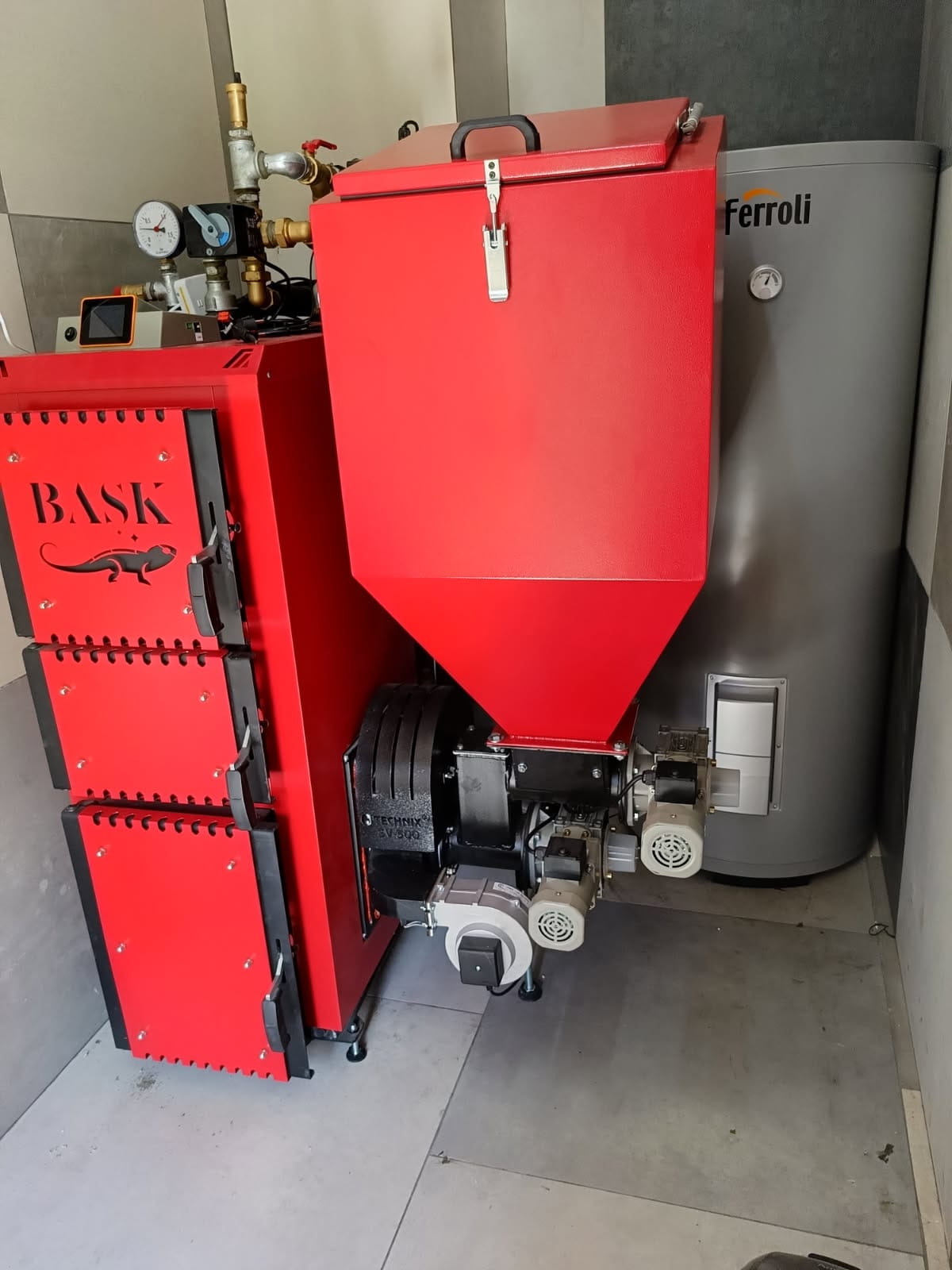 Czerwony piec z podajnikiem marki BASSK i szary zbiornik Ferolli w kotłowni. Widoczne manometry i zawory. Ujęcie z bliska, neutralne oświetlenie.