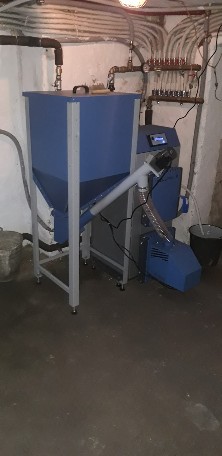 Niebiesko-szary piec na pellet z podajnikiem, widoczny w kotłowni z instalacją grzewczą na ścianie. Ujęcie z dołu, oświetlenie sufitowe.