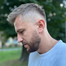 Skalski Pracownia Fryzur - Męska fryzura z cieniowaniem, widok z profilu. Krótkie, platynowe włosy z delikatnym zarostem. Naturalne światło, zieleń w tle.