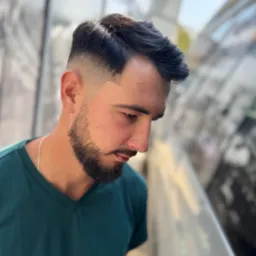 Męska fryzura z cieniowaniem i brodą, widok z góry, na zewnątrz, naturalne światło, model w zielonej koszulce, srebrny łańcuszek na szyi.