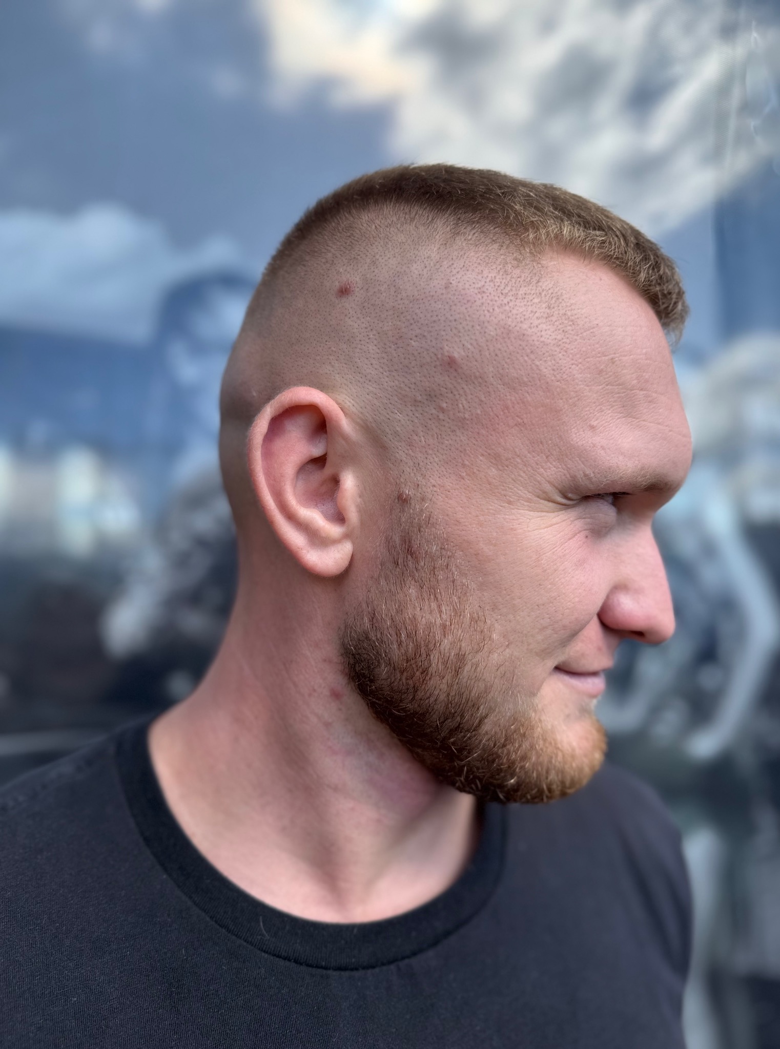 Mężczyzna z krótką fryzurą i zadbaną brodą, profil. Widoczne detale skóry głowy i zarostu. Tło rozmyte, przypomina abstrakcyjny wzór.