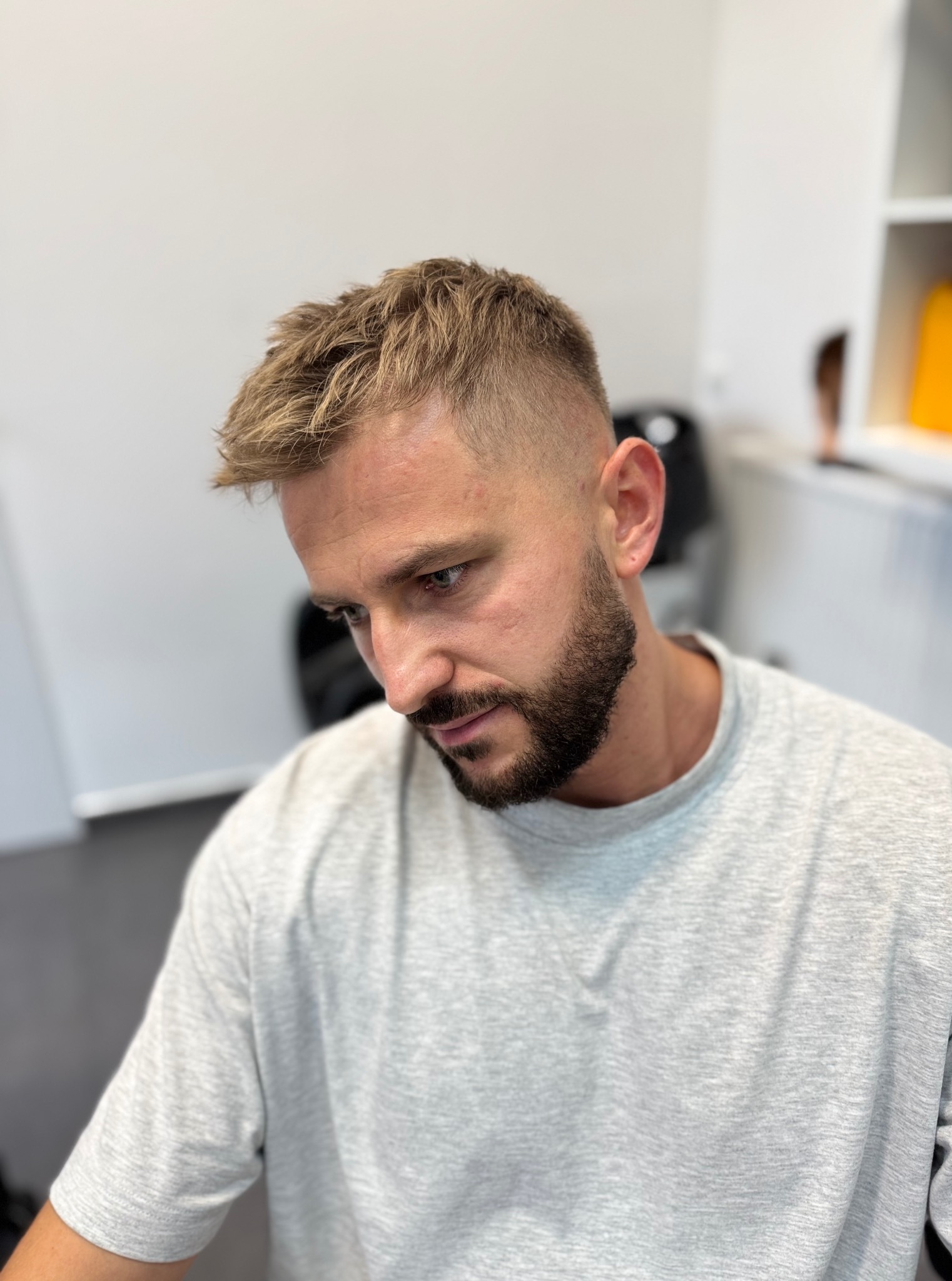 Męska fryzura z krótkimi bokami i dłuższą górą, zaczesana do góry, w odcieniach blondu. Widoczna zadbana broda. Mężczyzna patrzy w dół.