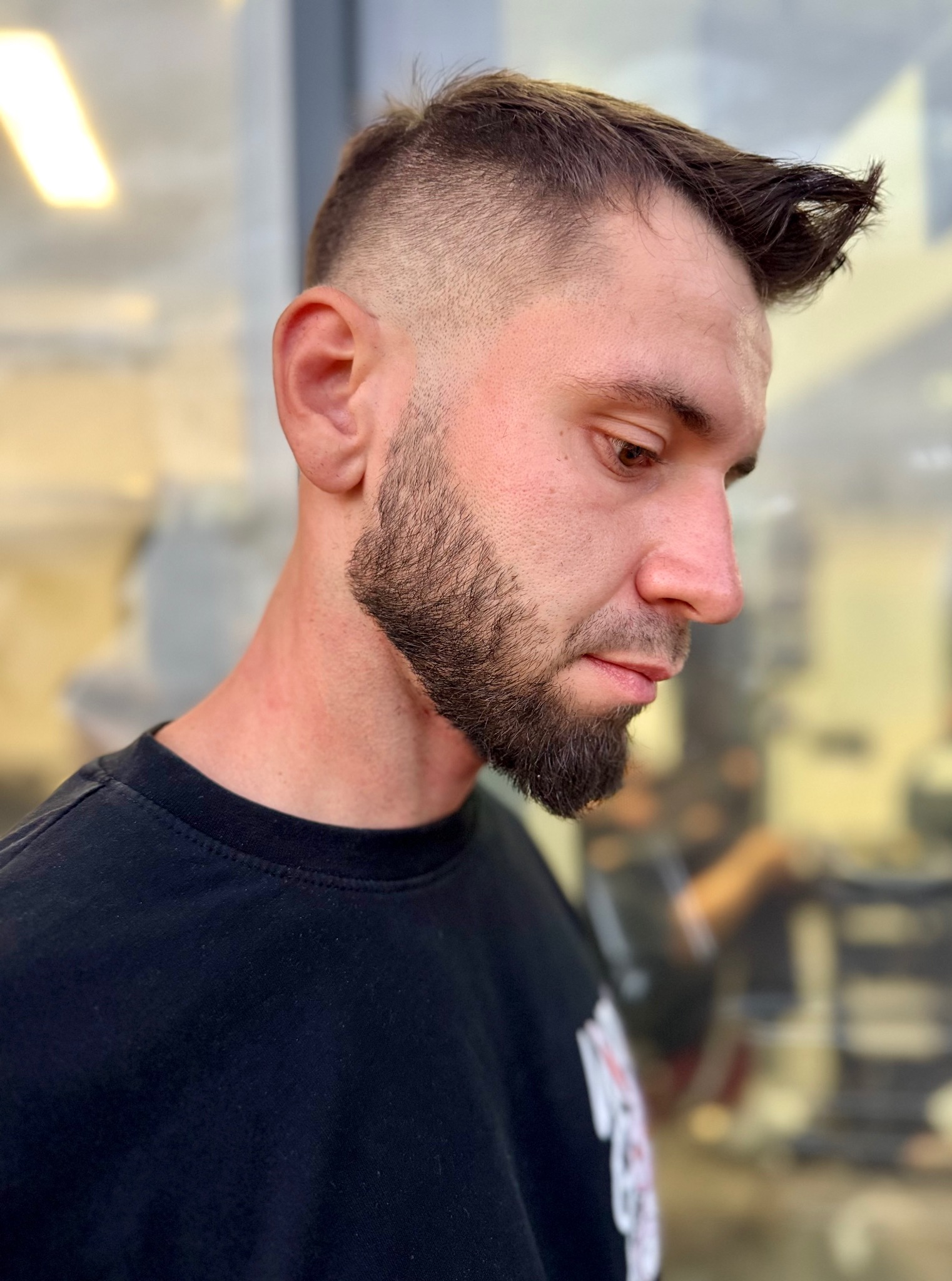 Mężczyzna z modną fryzurą undercut i starannie przystrzyżoną brodą, spoglądający w dół, w czarnej koszulce. Widoczne detale strzyżenia i cieniowania.