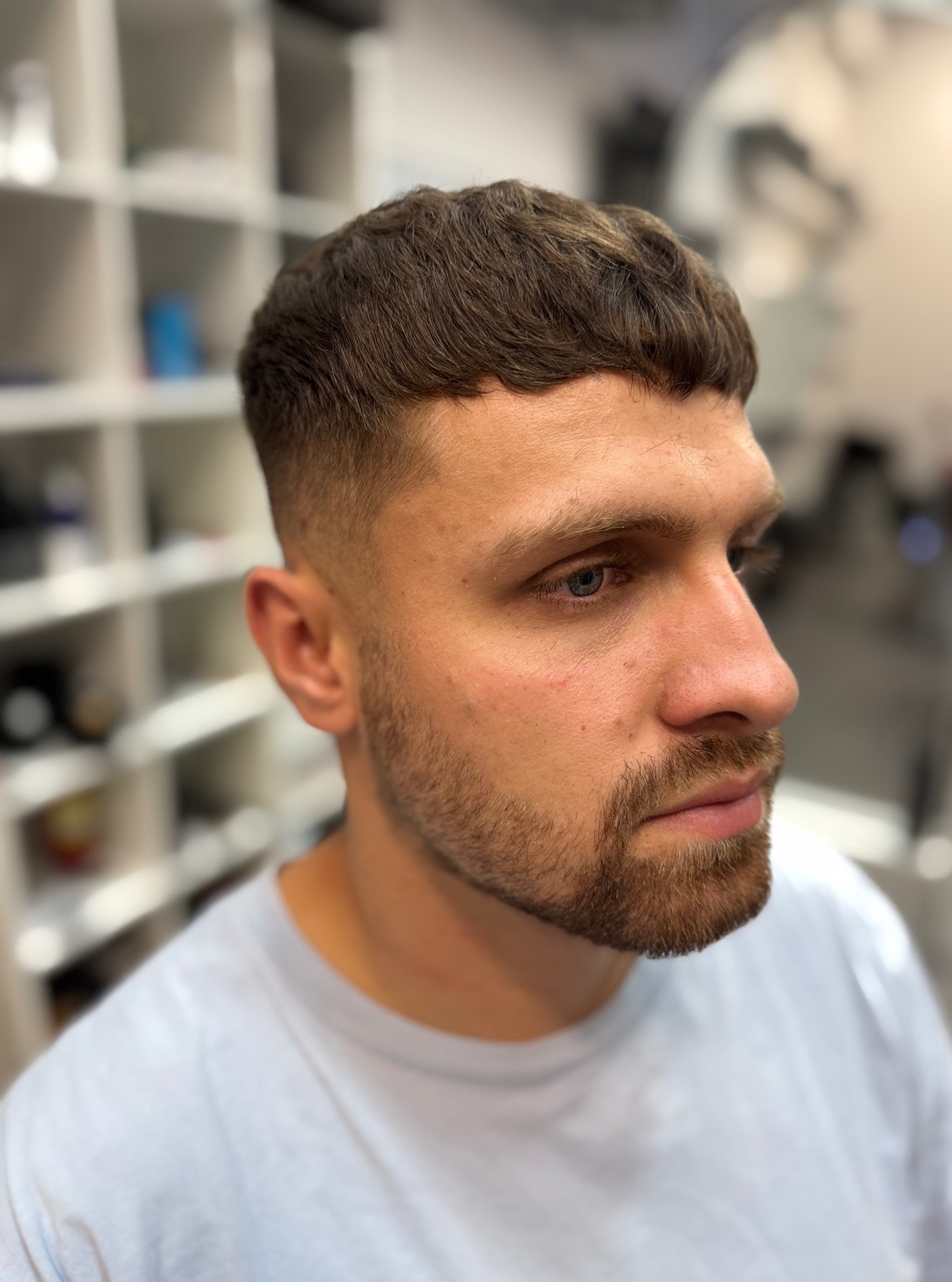 Męska fryzura z krótkim, równym cięciem grzywki i wycieniowanymi bokami, brązowe włosy, broda. Widok z profilu na tle białych półek.