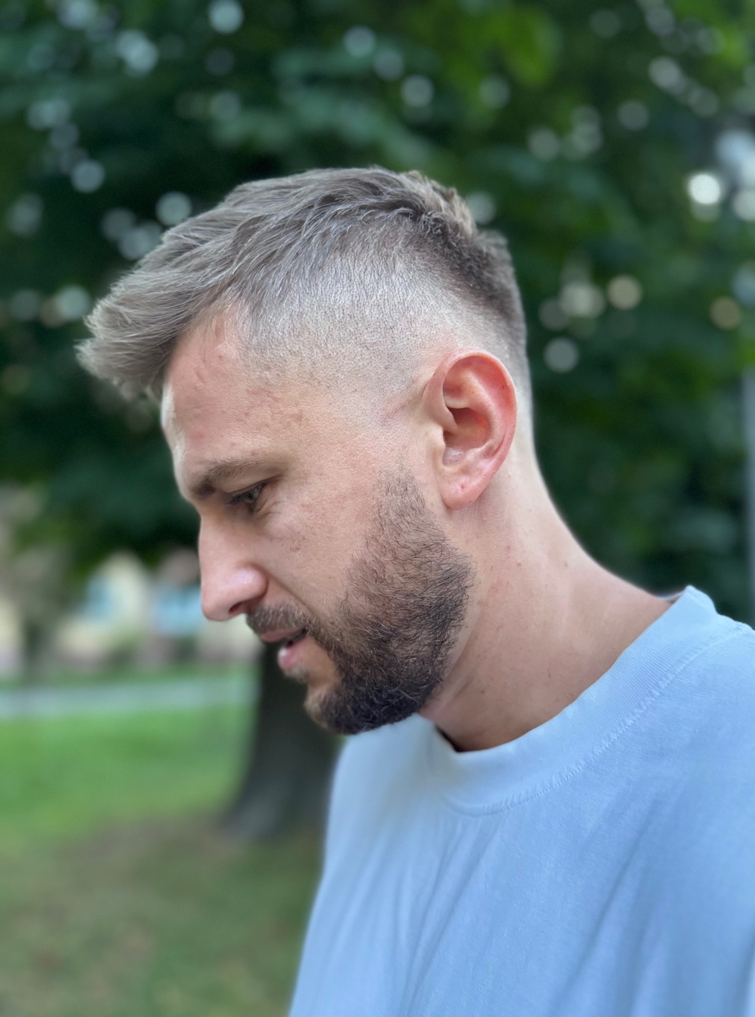 Męska fryzura z cieniowaniem, widok z profilu. Krótkie, platynowe włosy z delikatnym zarostem. Naturalne światło, zieleń w tle.