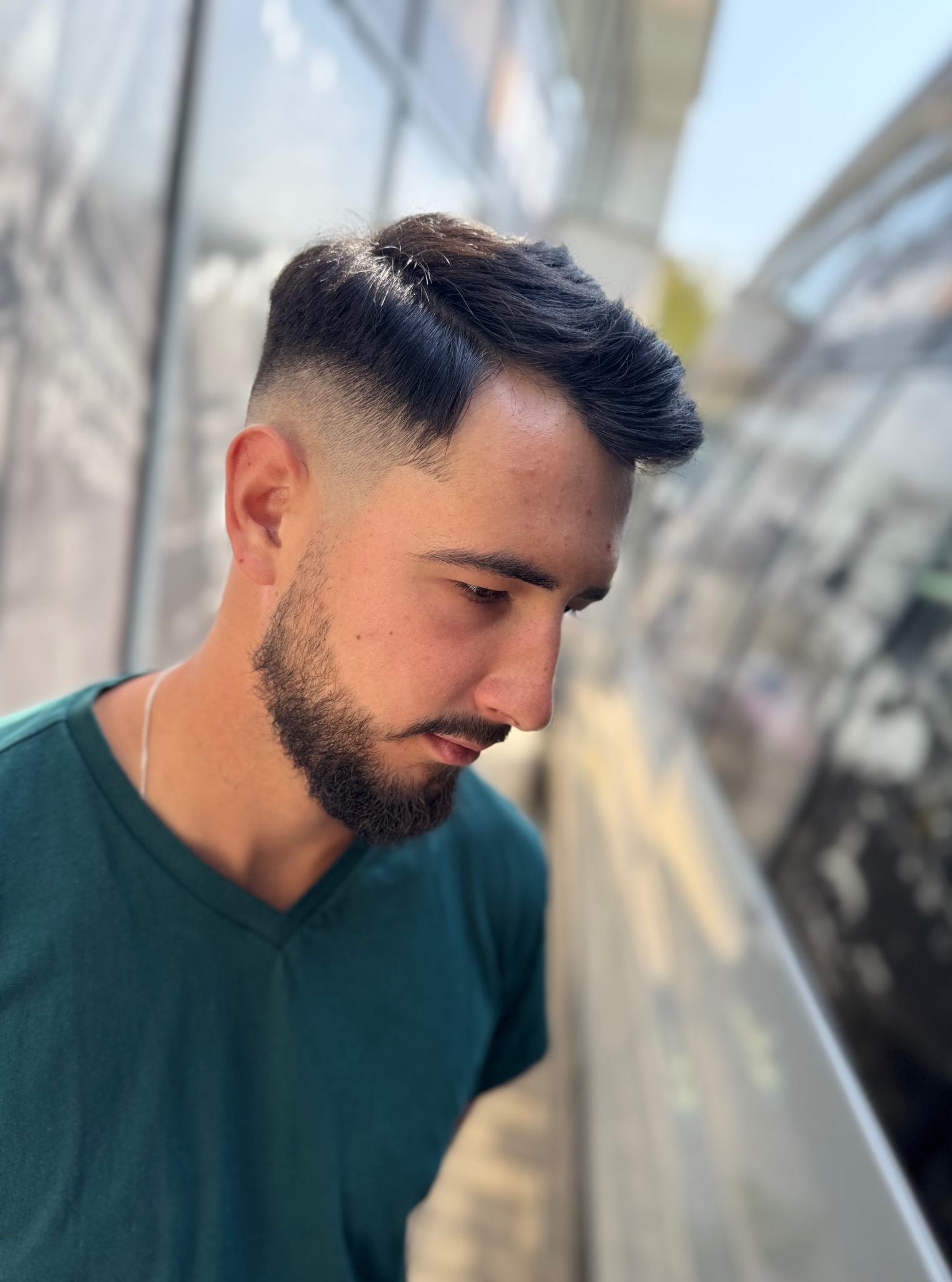 Męska fryzura z cieniowaniem i brodą, widok z góry, na zewnątrz, naturalne światło, model w zielonej koszulce, srebrny łańcuszek na szyi.