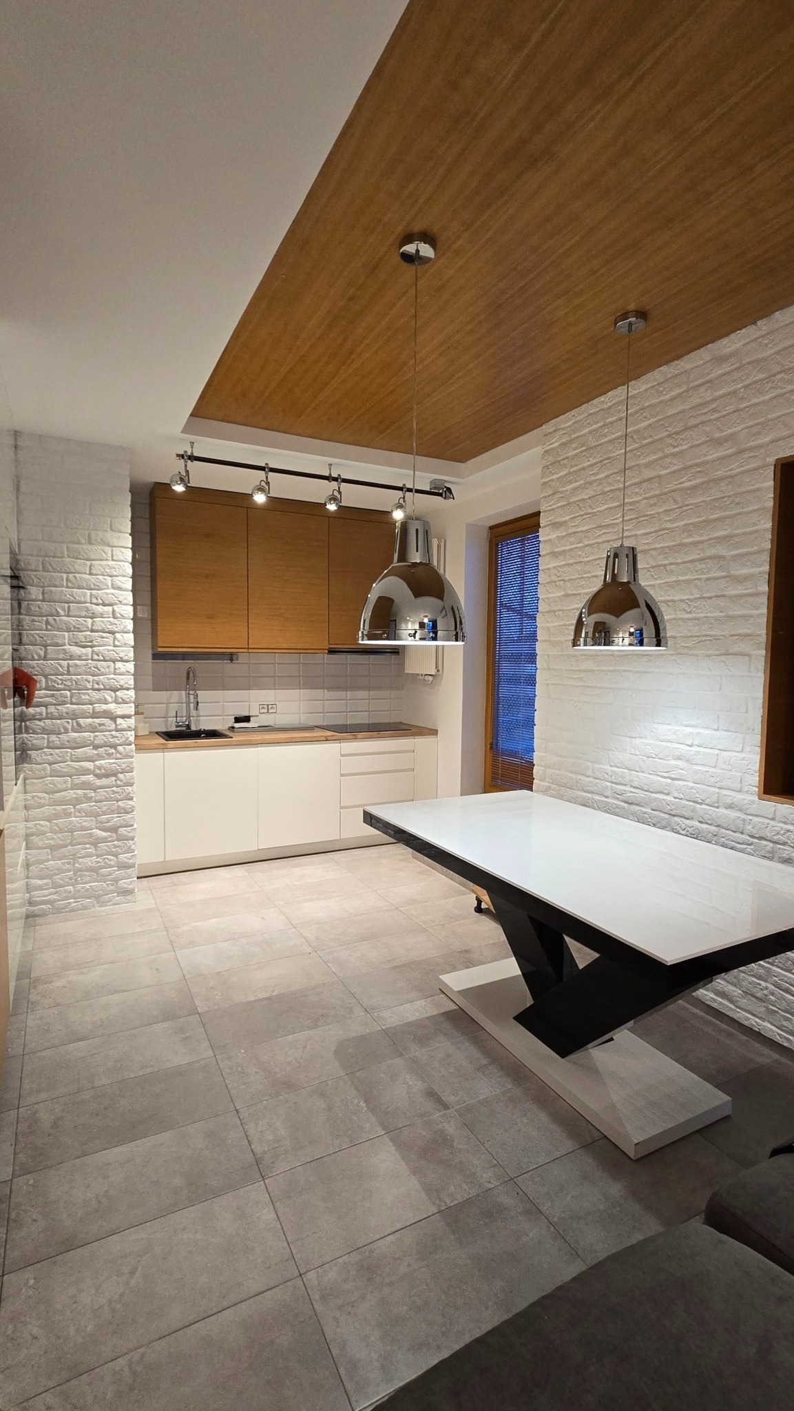 Nowoczesna kuchnia po wykończeniu w Rudzie Śląskiej: białe szafki, ceglana ściana, drewniany sufit i designerskie lampy wiszące nad stołem.