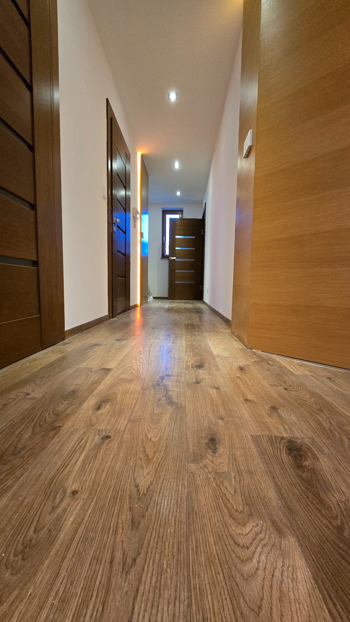 Nowoczesny korytarz z drewnianą podłogą i drzwiami w odcieniach brązu. Widok z niskiej perspektywy podkreśla głębię i wzór paneli. Oświetlenie punktowe w suficie.