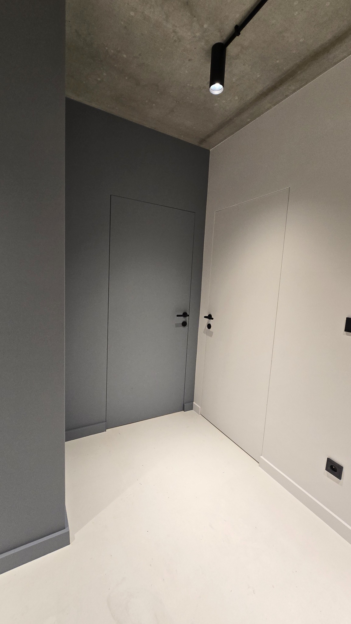 Minimalistyczne wnętrze: szara ściana z ukrytymi drzwiami obok białej ściany z drzwiami, kremowa podłoga, surowy betonowy sufit z czarnym reflektorem.