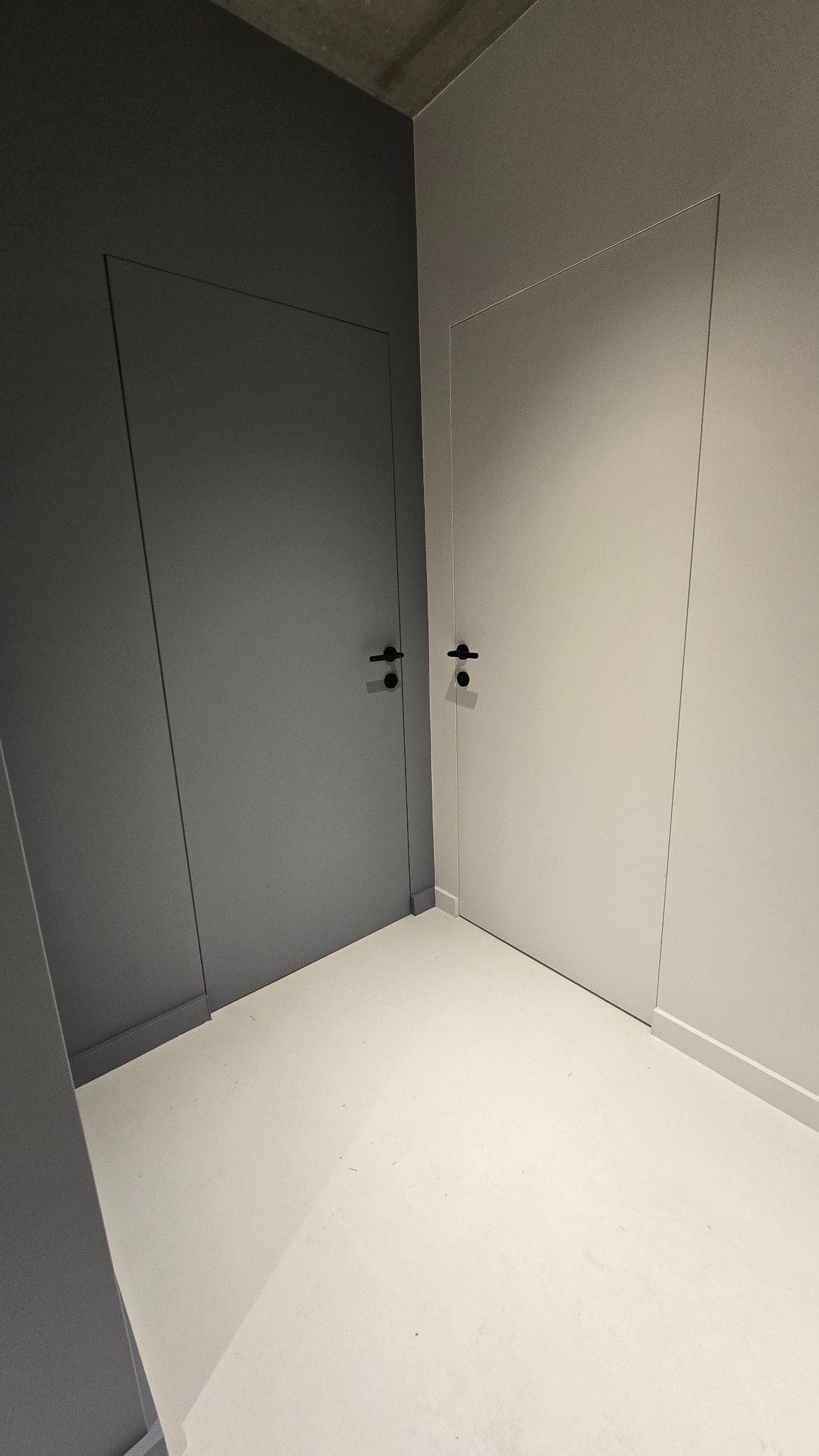 Dwie minimalistyczne, bezprzylgowe drzwi w odcieniach szarości i bieli, z czarnymi klamkami, w jasnym wnętrzu. Kontrast kolorów podkreśla nowoczesny design.