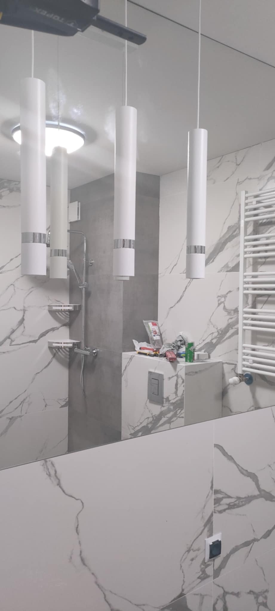 Wykończona łazienka z marmurowymi płytkami, białymi lampami wiszącymi i grzejnikiem drabinkowym. Widoczne narzędzia i akcesoria na blacie.