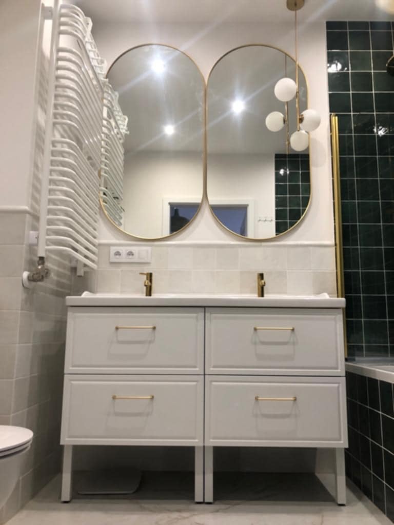 Nowoczesna łazienka z podwójną umywalką, owalnymi lustrami w złotych ramach i grzejnikiem drabinkowym. Białe płytki i armatura tworzą elegancki wystrój.