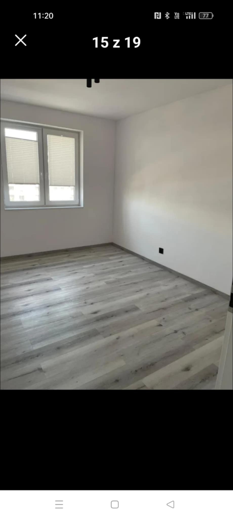 Jasne panele podłogowe w pokoju z białymi ścianami i oknem z żaluzjami plisowanymi. Minimalistyczne wnętrze po remoncie, widok na podłogę i fragment ściany.