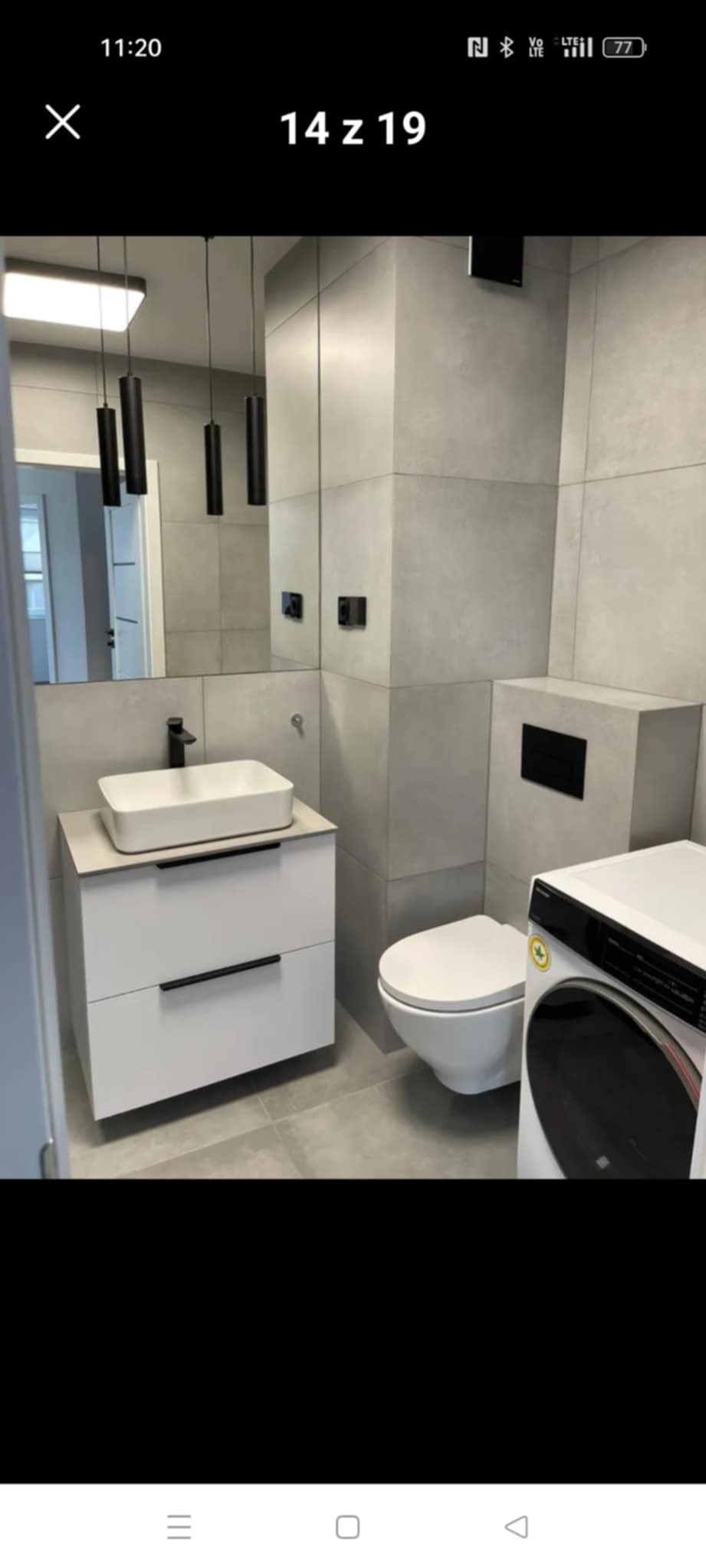 Nowoczesna łazienka z szarymi płytkami, białą umywalką na szafce z szufladami i wiszącą toaletą. Minimalistyczne oświetlenie i pralka w tle.