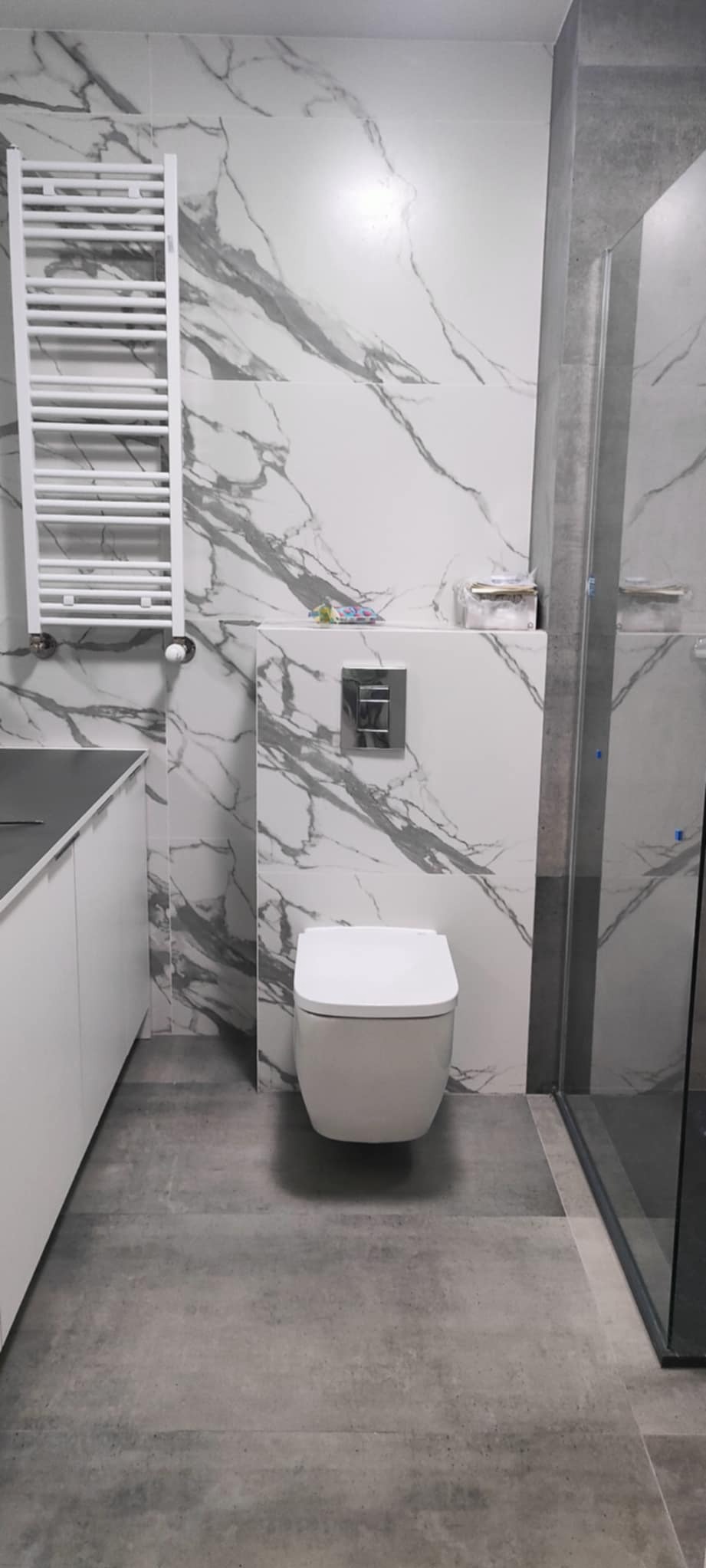 Nowoczesna łazienka z marmurowymi płytkami, podwieszaną toaletą i grzejnikiem drabinkowym. Szare płytki podłogowe i minimalistyczny design.