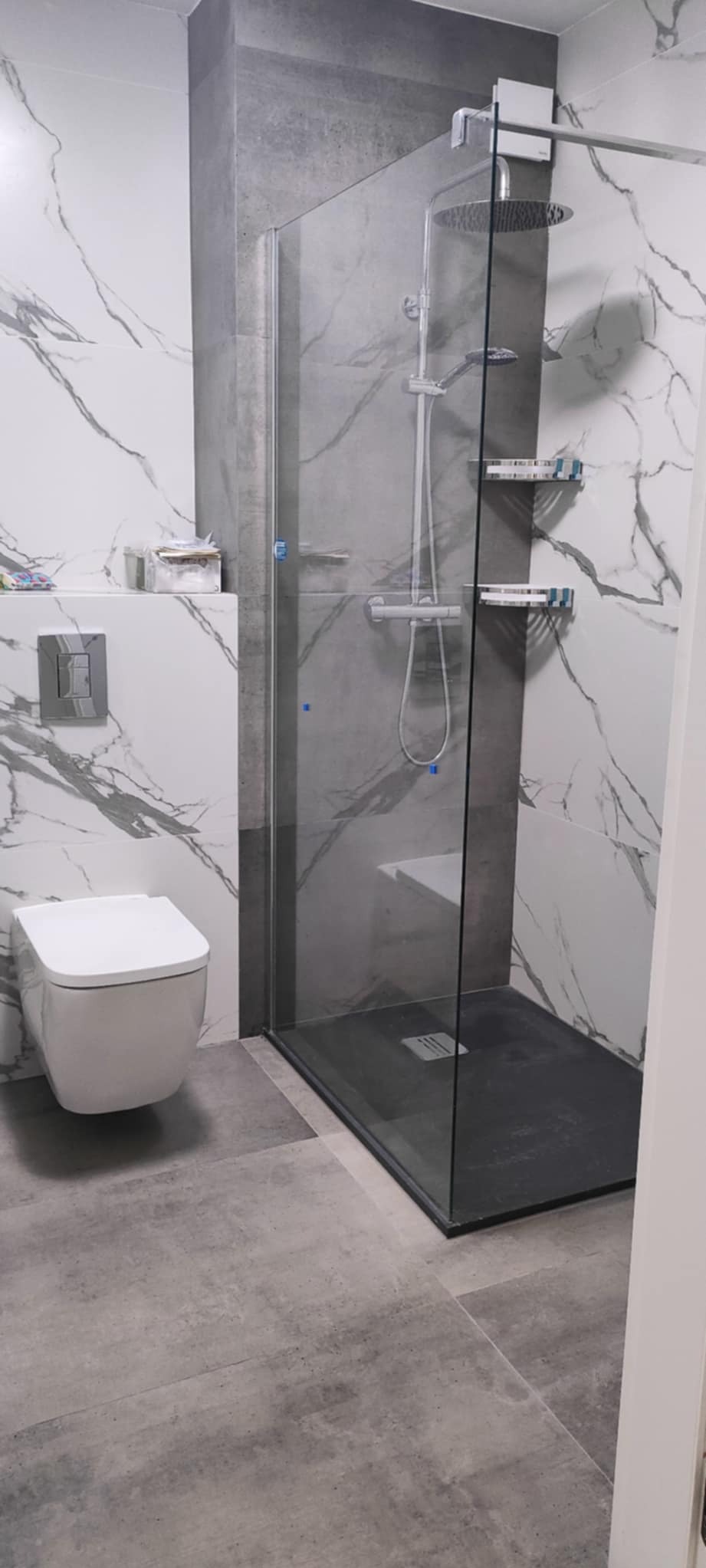 Nowoczesna łazienka z kabiną prysznicową typu walk-in, wykończona płytkami imitującymi marmur i beton. Minimalistyczna toaleta podwieszana i szara podłoga.