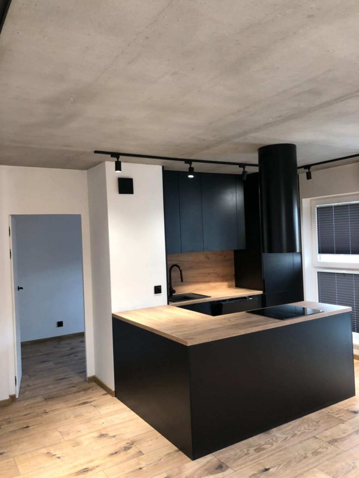 Nowoczesna kuchnia z czarnymi szafkami i drewnianym blatem, oświetlenie punktowe, minimalistyczny design, widok na parkiet i otwarte przejście do innego pomieszczenia.