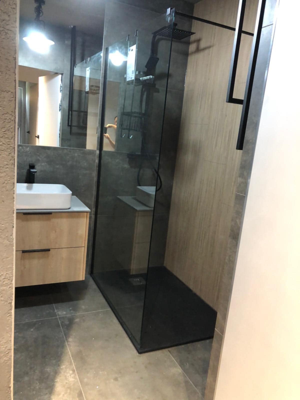 Nowoczesna łazienka z kabiną typu walk-in, umywalką nablatową na szafce z drewnopodobnym frontem i szarą armaturą. Minimalistyczny design, szare płytki i oświetlenie.