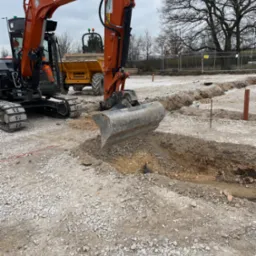 Pomarańczowa koparka HACS w trakcie wykopu wąskiego rowu na żwirowym terenie budowy. W tle żółta wywrotka Thwaites i częściowo widoczne drzewa.