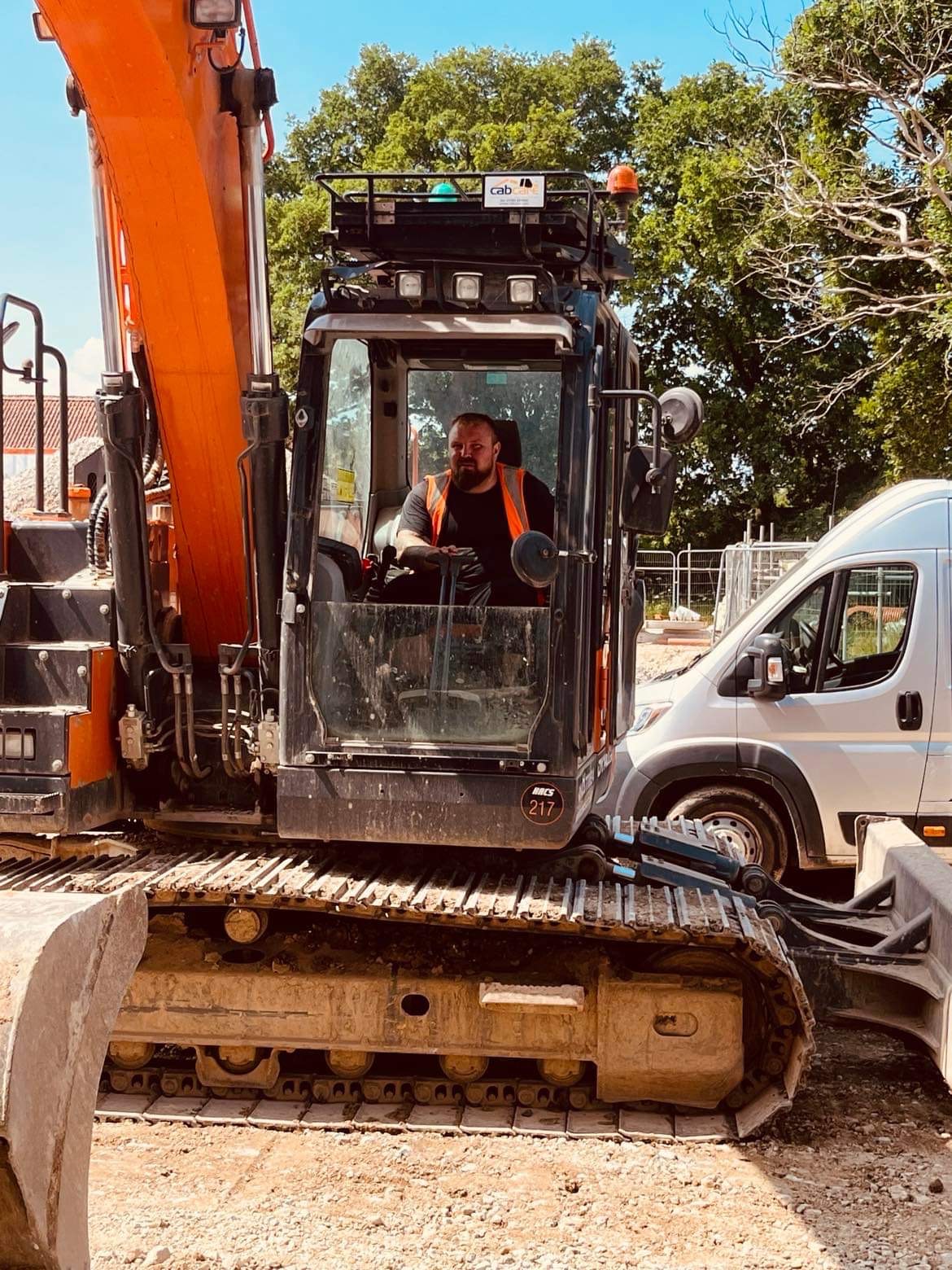Operator w pomarańczowej kamizelce siedzi w kabinie pomarańczowo-czarnej koparki, obok stoi biały van. W tle zieleń drzew i częściowo widoczny budynek.