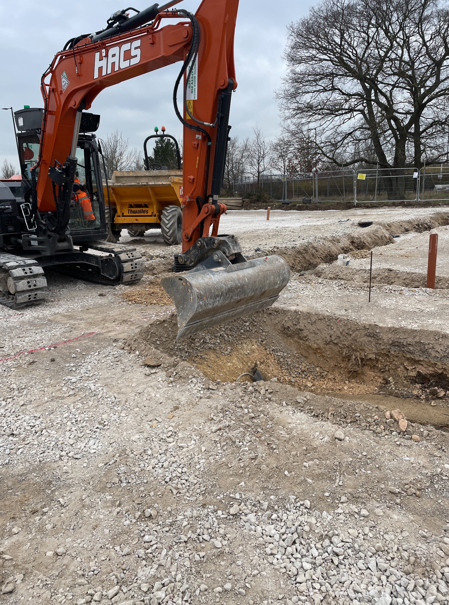 Pomarańczowa koparka HACS w trakcie wykopu wąskiego rowu na żwirowym terenie budowy. W tle żółta wywrotka Thwaites i częściowo widoczne drzewa.