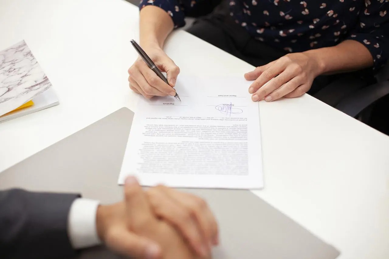 Osoba podpisuje dokument na białym biurku, widoczne dłonie, długopis i fragment garnituru drugiej osoby. Umowa z niebieskim podpisem.