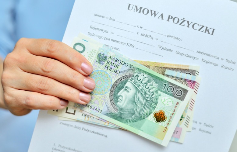 Umowa pożyczki trzymana w dłoni z banknotami 100 zł. Dokument z danymi rejestrowymi firmy. Kredyt gotówkowy i formalności, w tle błękitna koszula.