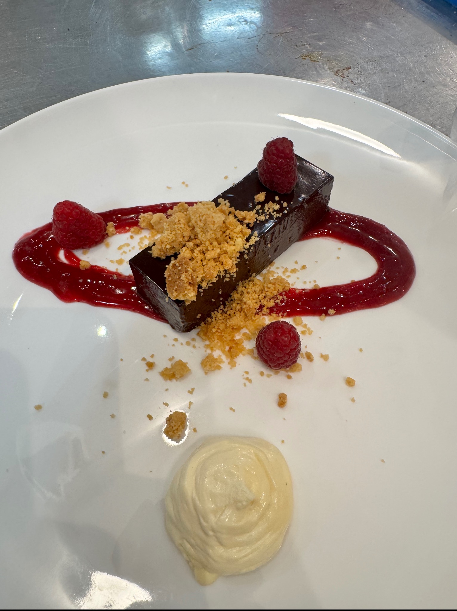 Elegancko podany deser: czekoladowy blok z kruszonką, malinami i sosem owocowym na białym talerzu. Minimalistyczna kompozycja z kremowym akcentem.