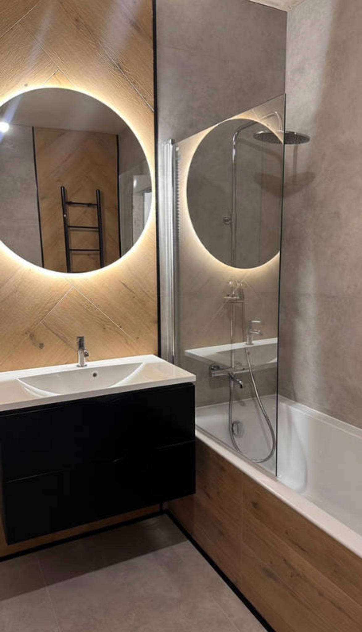 Nowoczesna łazienka z okrągłym lustrem podświetlanym LED i drewnopodobnymi płytkami w Poznaniu. Umywalka z czarną szafką, szklany parawan nawannowy.