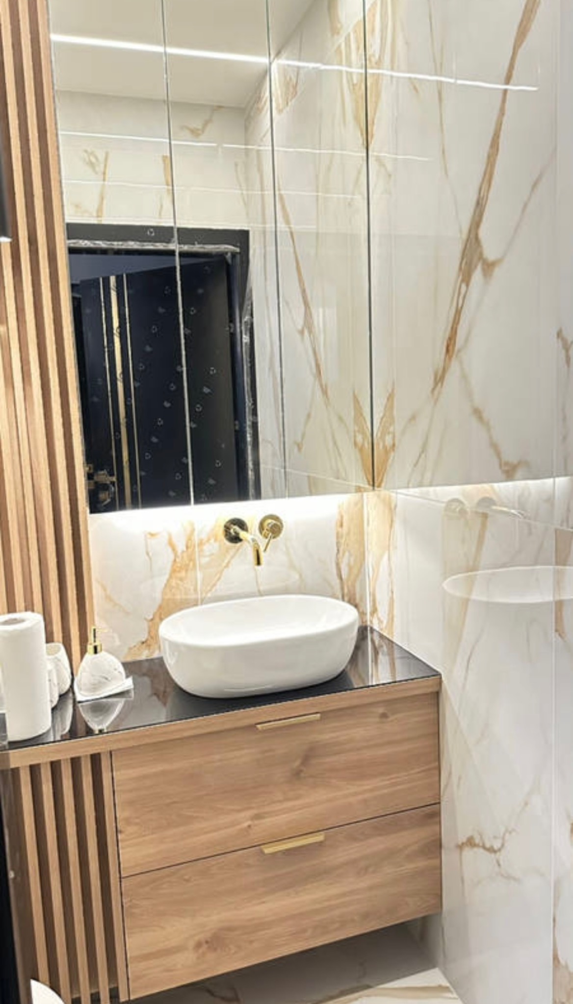 Nowoczesna łazienka z marmurowymi płytkami, drewnianą szafką pod umywalką i złotą armaturą. W lustrze odbija się czarne drzwi.