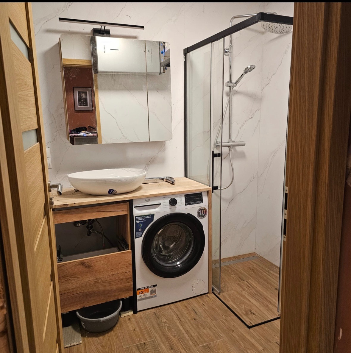 Nowoczesna łazienka z kabiną prysznicową, umywalką nablatową i pralką zabudowaną w szafce. Widoczne białe płytki imitujące marmur i drewniane akcenty wykończeniowe.