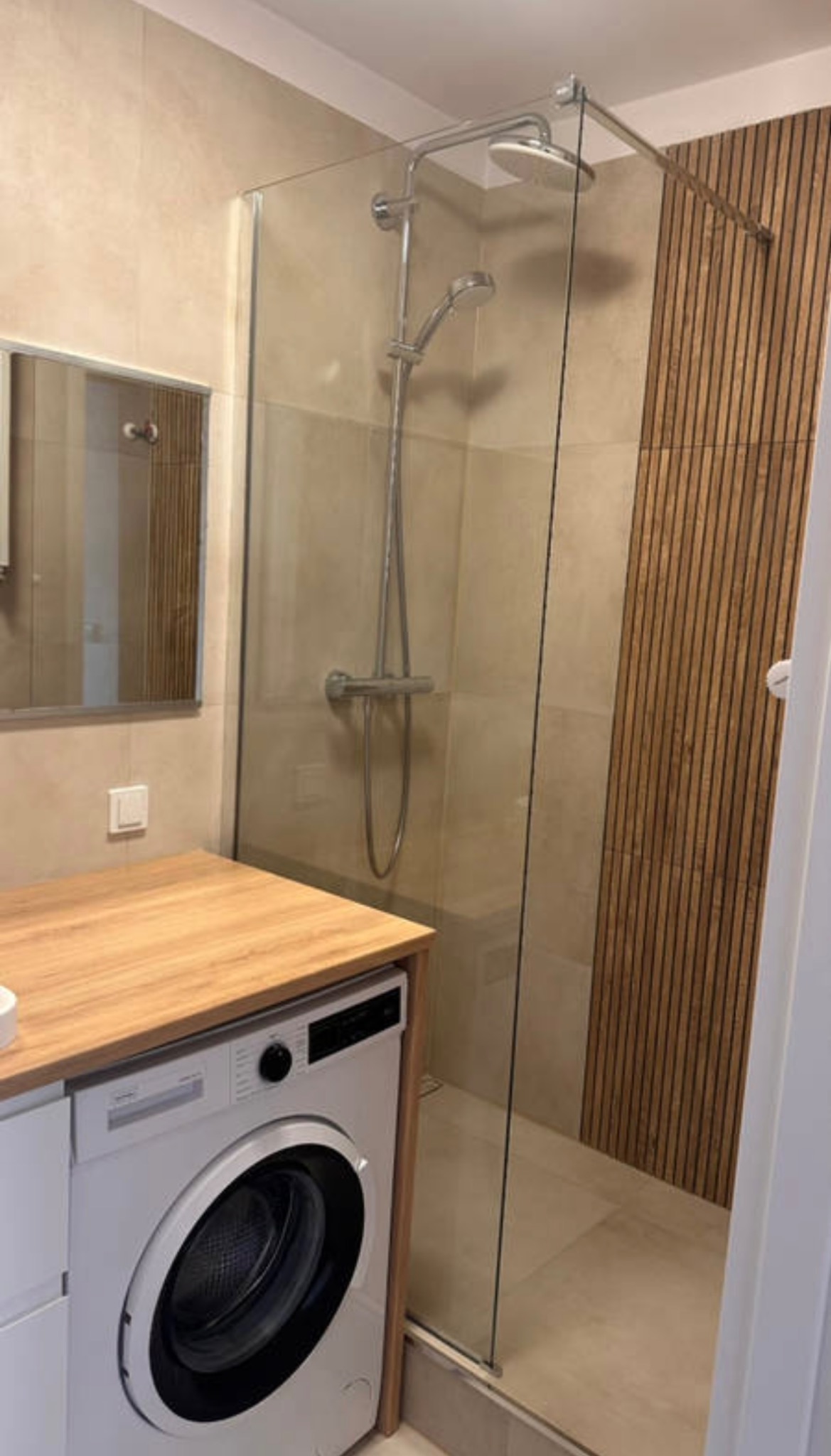 Nowoczesna łazienka z kabiną prysznicową typu walk-in, drewnianymi panelami ściennymi i pralką pod blatem. Minimalistyczny design z beżową kolorystyką.