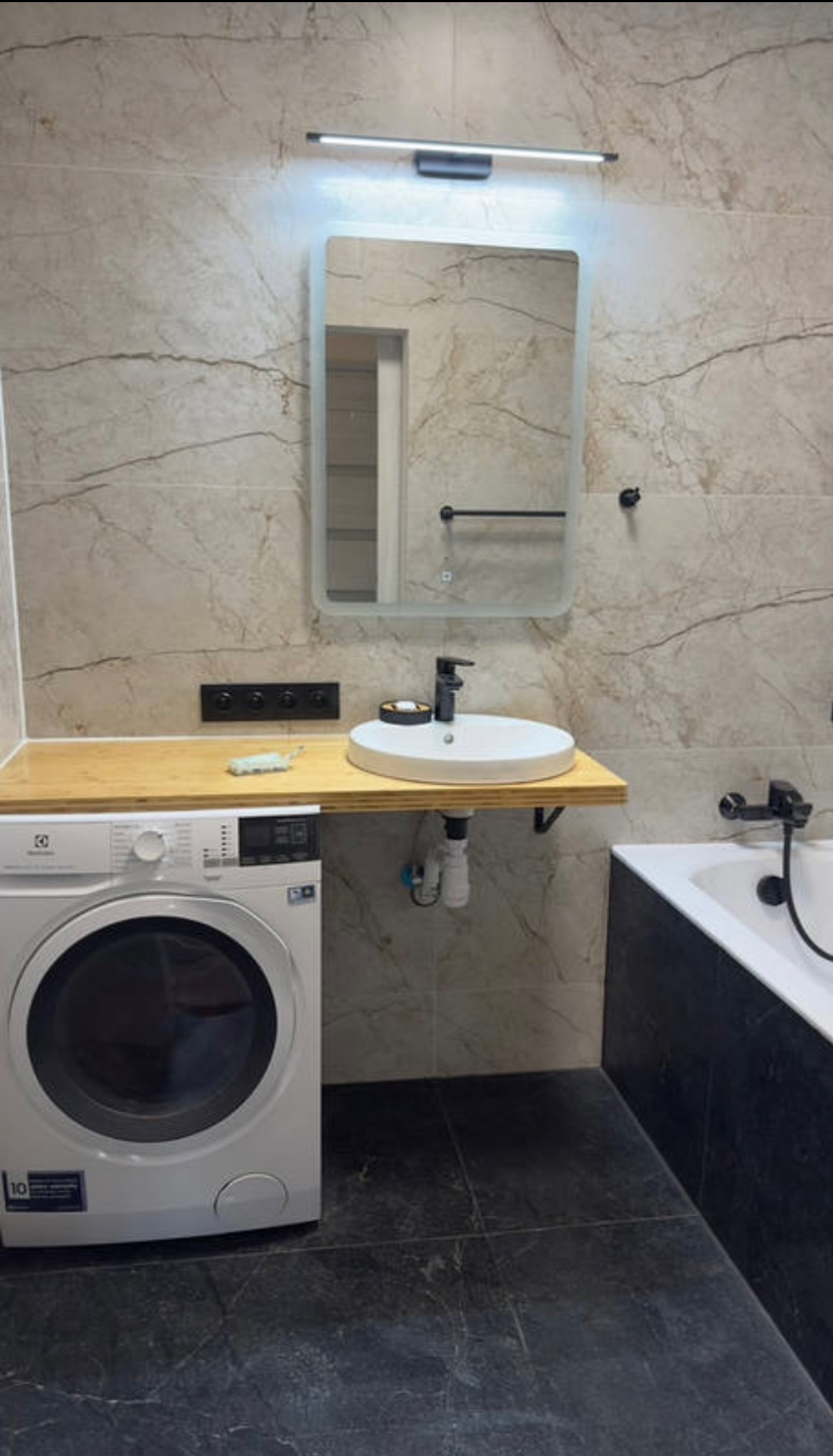 Nowoczesna łazienka po remoncie: biała umywalka nablatowa, lustro z podświetleniem LED, pralka i marmurowe płytki ścienne w odcieniach beżu i szarości.